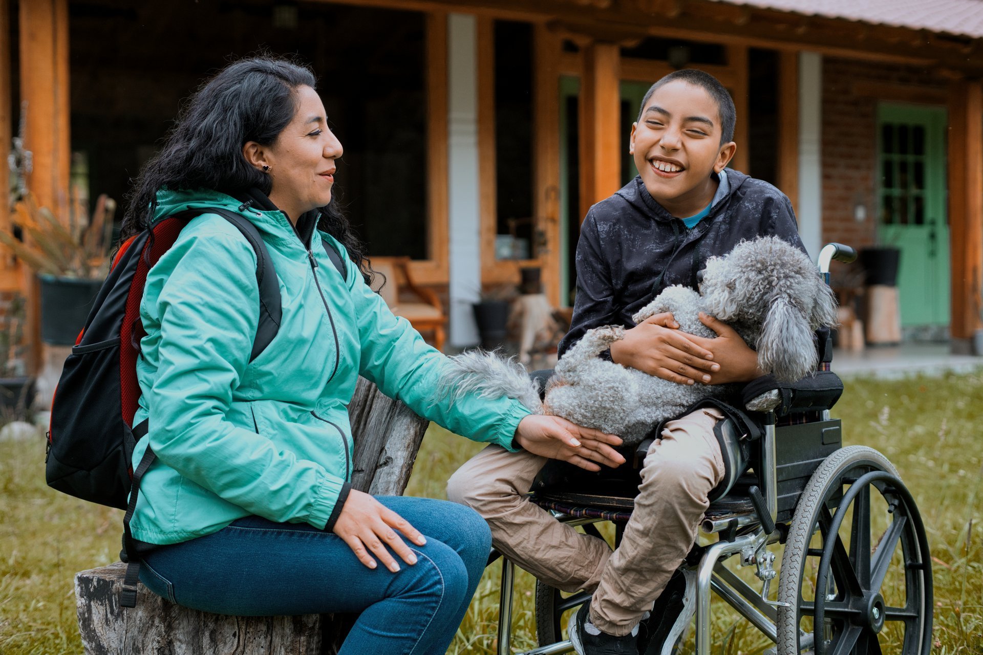 Joyful boy in a wheelchair holding a fluffy dog while sitting outdoors with a woman, sharing a moment of happiness